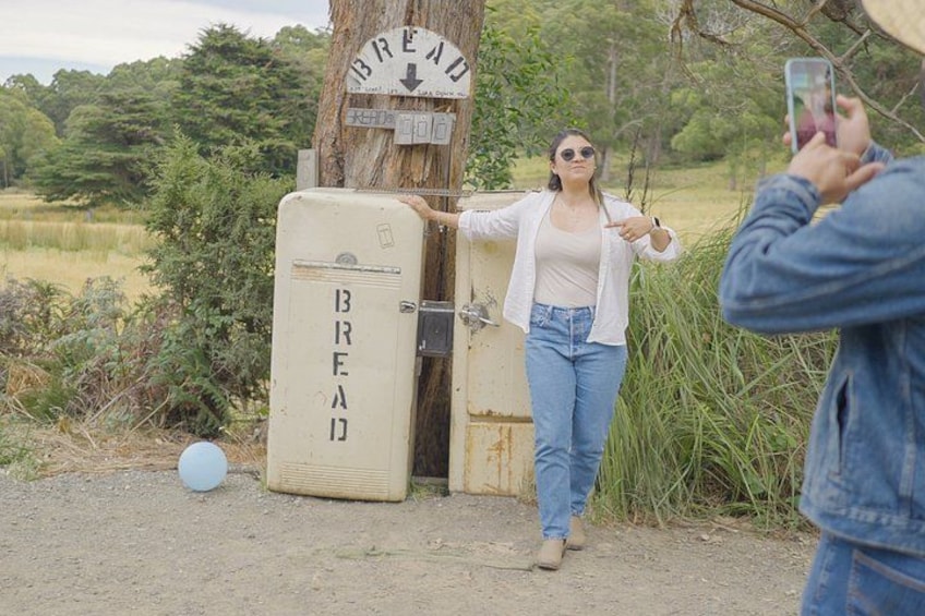 A woman standing next to a vintage refrigerator with 'BREAD' written on it, a tree, and a person taking a photo. 2