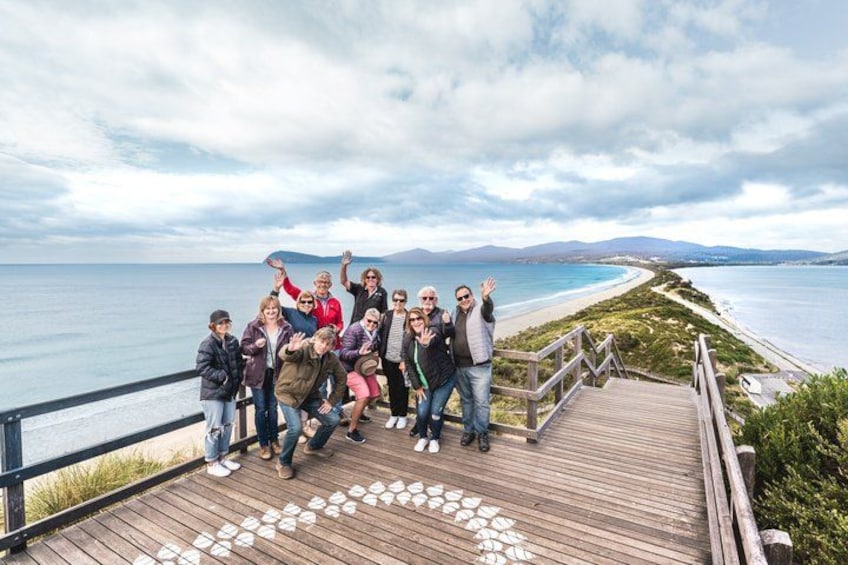 Un grupo de personas en una pasarela de madera contemplando una playa con vistas claras al océano y montañas distantes. 1