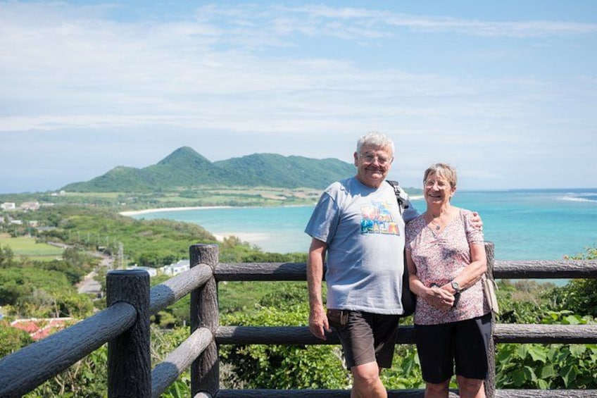A couple standing on a wooden railing overlooking a coastal landscape with mountains and a beach. 1