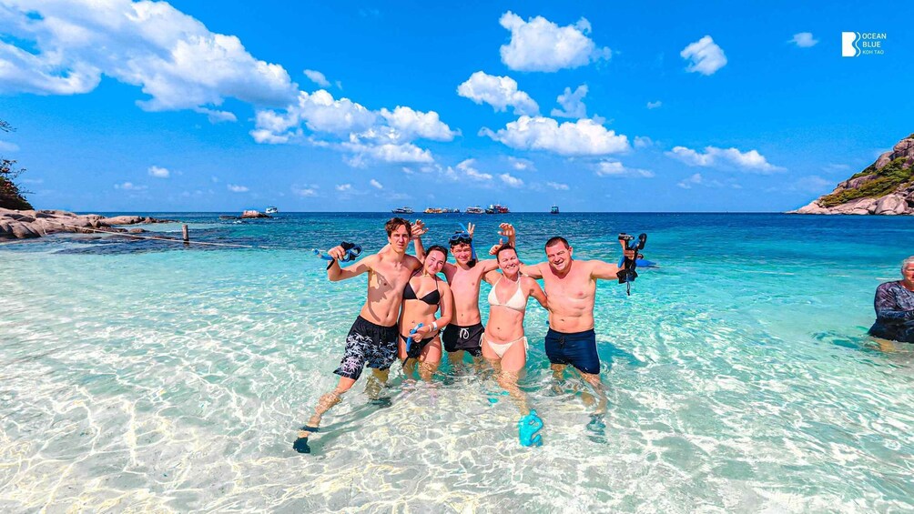 A group of people in a clear blue sea with rocky shores and boats in the distance. 4