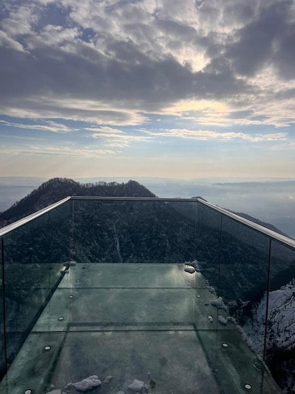 A glass-enclosed observation deck with a view of a forested mountain and a cloudy sky. 4