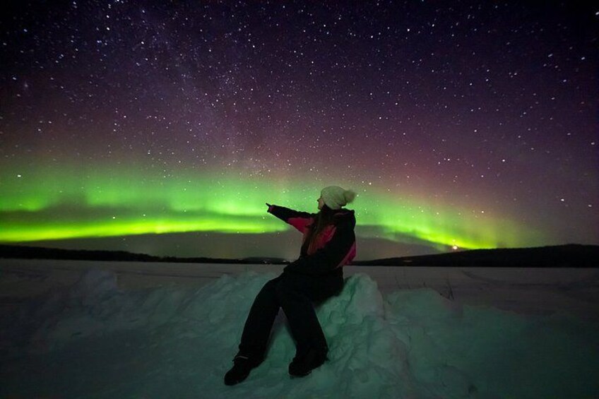 Une personne assise sur une surface recouverte de neige, contemplant le ciel nocturne où l’on peut voir des étoiles et des aurores boréales. 3