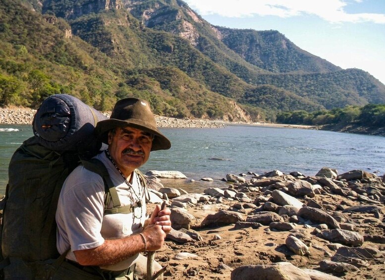 A person with a backpack standing by a river with rocky banks and mountains in the background. 4