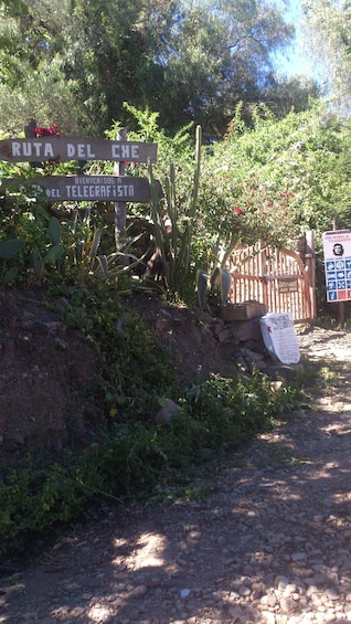 A signpost with 'RUTA DEL CHE' and 'BIENVENIDOS A TELEGRAFISTA' written on it, surrounded by greenery and a stone wall. 3