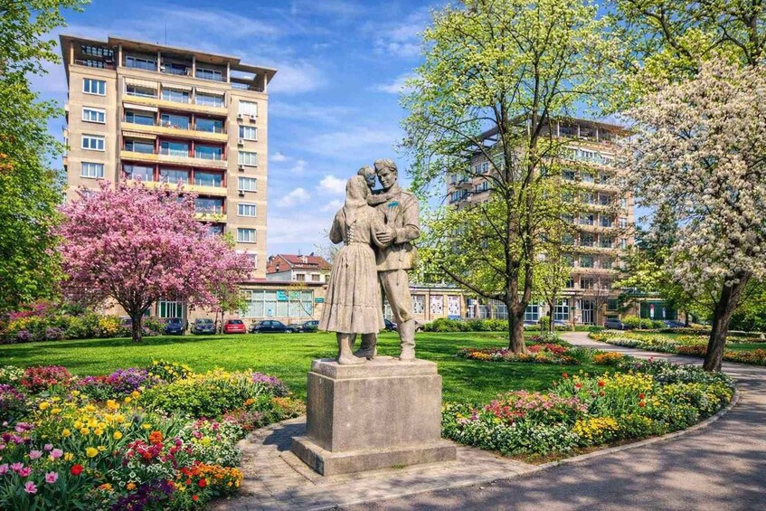 A statue of a couple in a park with flowering trees and a multi-story building in the background. 1