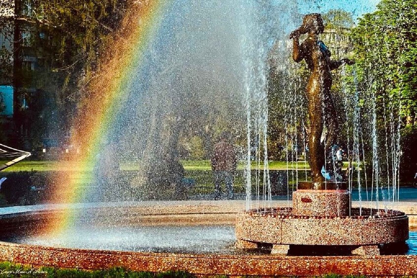 A fountain with water jets and a statue, surrounded by a circular stone structure, with a rainbow in the background. 2