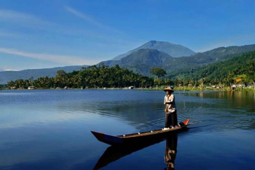 A calm lake with a person standing in a boat, surrounded by lush greenery and mountains in the background. 1