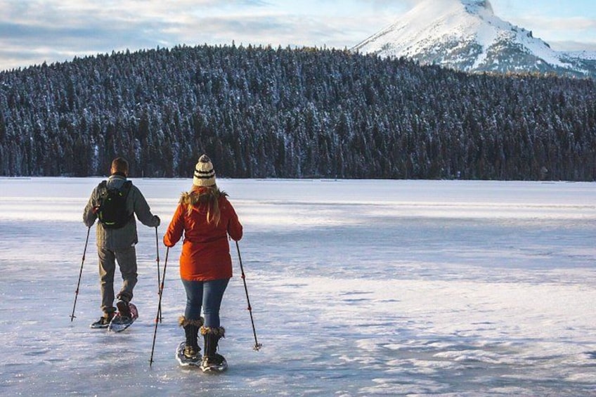Dos personas practican esquí de fondo en un lago congelado, con un bosque y una montaña nevada al fondo. 1