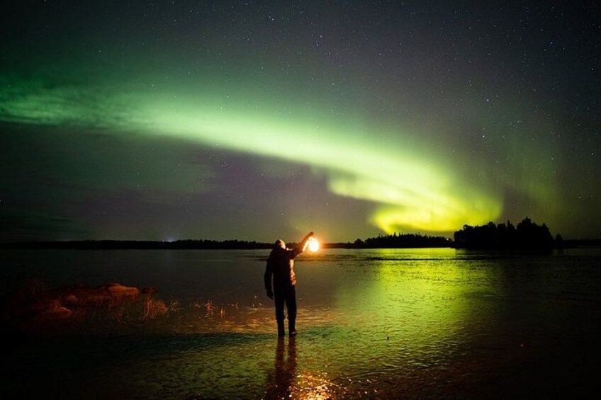 Iemand staat bij een waterpartij in de nacht, met het Noorderlicht aan de hemel. 1