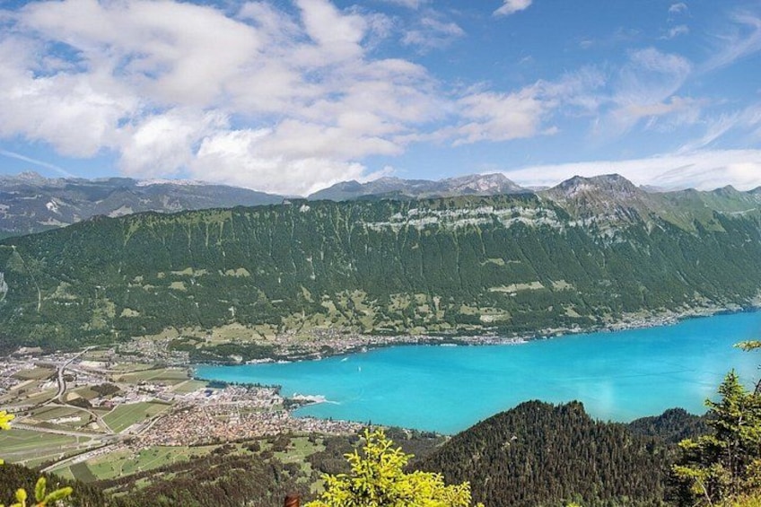 A coastal landscape with a turquoise bay, dense forested mountains, and a small town by the water. 5