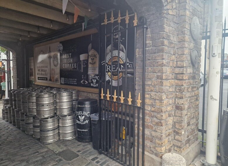 A brick building with a black gate, barrels, and a sign advertising Irish coffee and whiskey. 4