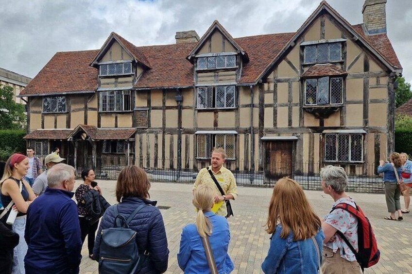 Eine Gruppe von Menschen steht vor einem historischen Gebäude im Tudorstil mit einer Ziegelaußenwand und zahlreichen Fenstern. 4