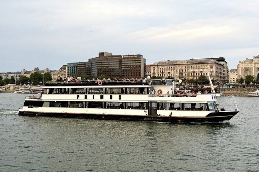Un bateau de croisière fluviale nommé PRIMUS, avec des passagers sur le pont, navigue devant un paysage urbain où se dressent des bâtiments historiques. 3