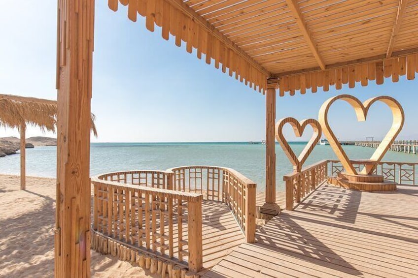Une terrasse en bois donnant sur la plage, décorée de motifs en forme de cœur et abritée par un parasol en chaume. 2