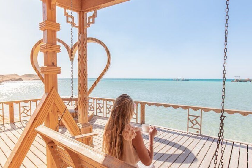 Une terrasse en bois avec une balançoire, d’où l’on peut admirer une mer bleue et limpide, avec des bateaux qui se dessinent à l’horizon. 4