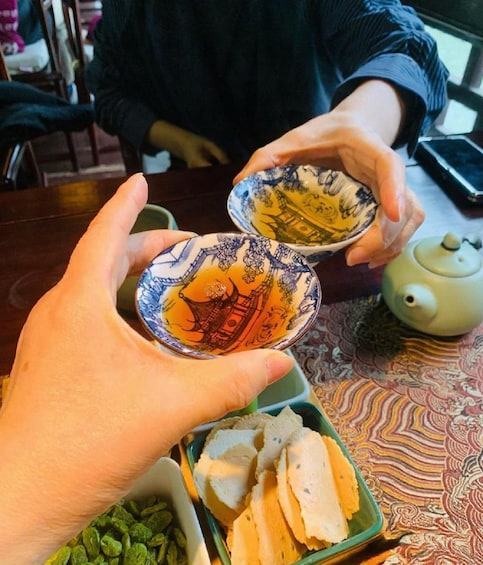 Two hands holding a pair of ornate bowls with traditional designs, a teapot, and a tray with food items. 2