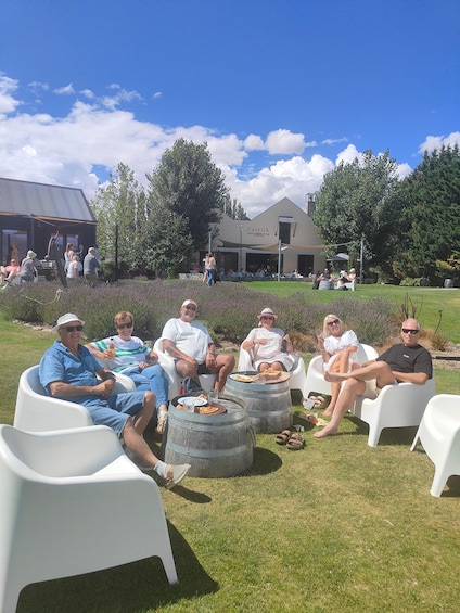 Een groep mensen zit buiten op witte stoelen, met eten en drinken, met een gebouw met de tekst 'Carrick' op de achtergrond en een gazon met bomen. 4