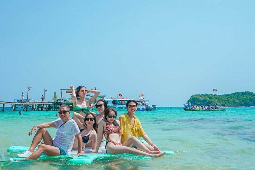 Un grupo de personas sentadas sobre una tabla de surf en agua turquesa cristalina, con un muelle y embarcaciones al fondo. 3