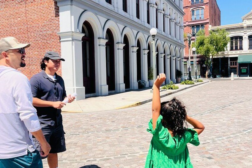 A person in a green dress standing in a plaza with a building featuring arched windows in the background. 1
