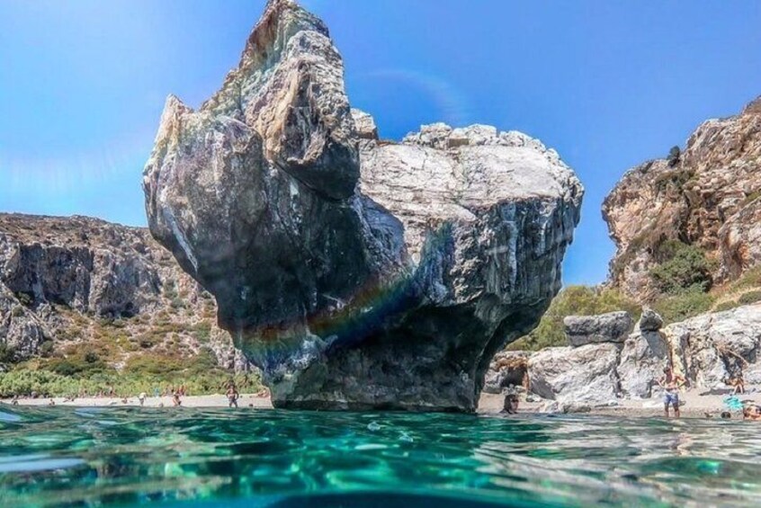 A large rock formation in clear water, surrounded by rocky cliffs and a few people in the background. 5