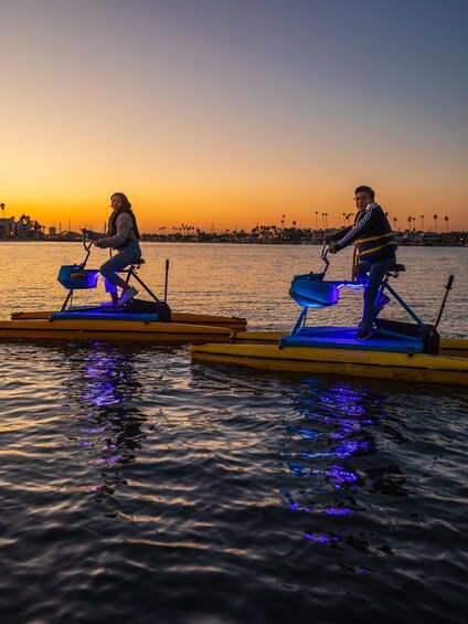 Two people riding stationary bikes on water platforms during sunset. 1