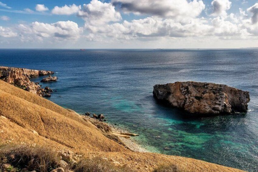 A coastal landscape with a prominent rocky outcrop in the sea, a clear blue sky, and a dry, grassy hillside. 5