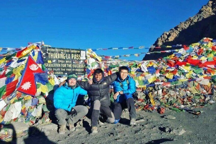 Three people sitting in front of a pile of colorful prayer flags with a sign that reads 'Thorang La Pass.' 5