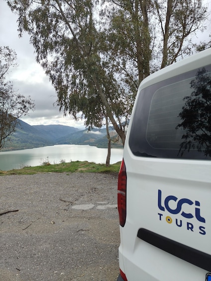 A white van with 'LOCI TOURS' on the back parked by a lake with mountains in the background. 5