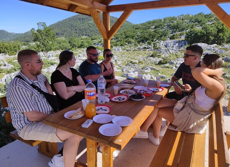 A group of people dining outdoors at a wooden table with plates, bottles, and food. 3