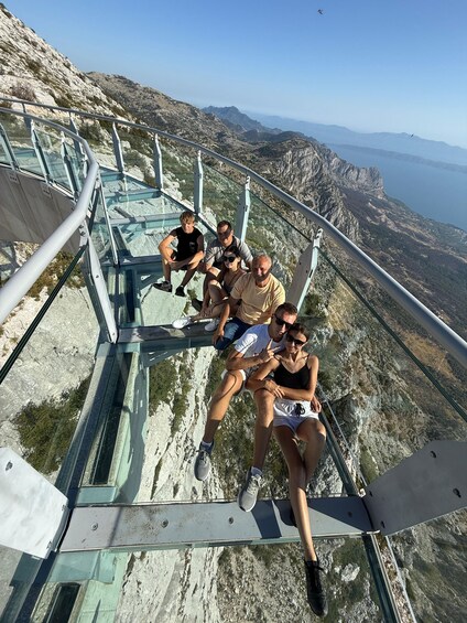 A group of people sitting on a glass bridge with a mountainous landscape in the background. 2