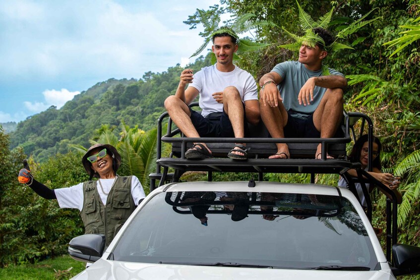 Three people on a roof rack of a white vehicle, surrounded by lush greenery and hills. 1