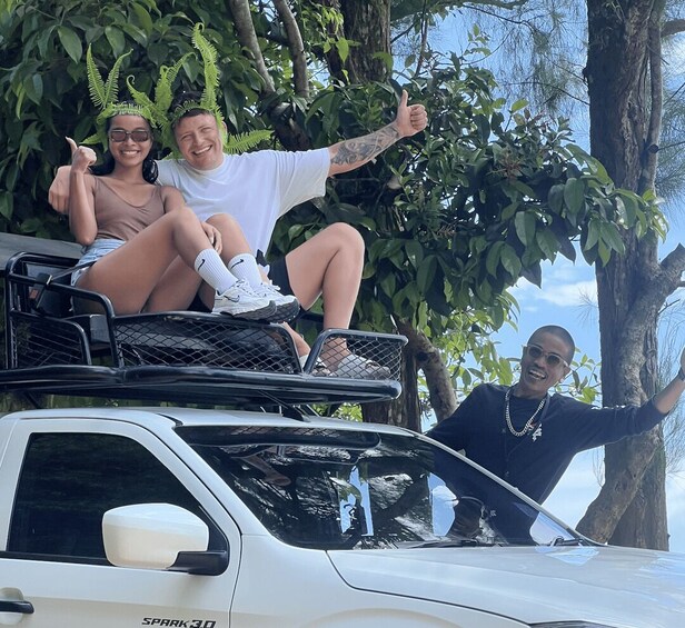 A white car with a roof rack, a person in a white shirt and shorts sitting on the roof rack, and another person in a black shirt standing beside the car. 4
