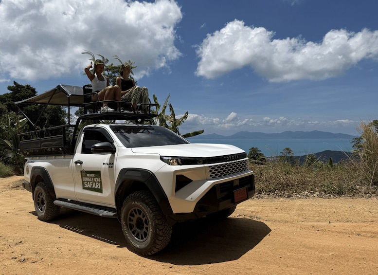 A white Toyota Hilux 4x4 with a roof rack, parked on a dirt road with a scenic view of mountains and a body of water in the background. 1