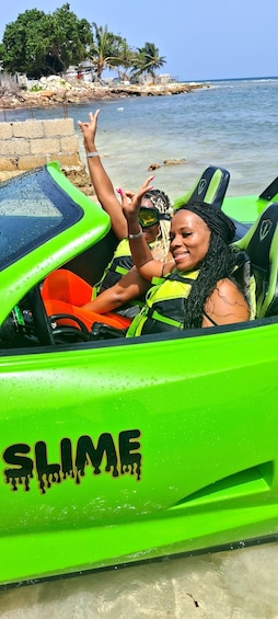 Dos personas en una embarcación verde con la palabra 'SLIME' en el costado, cerca de una playa con palmeras y un litoral rocoso. 2