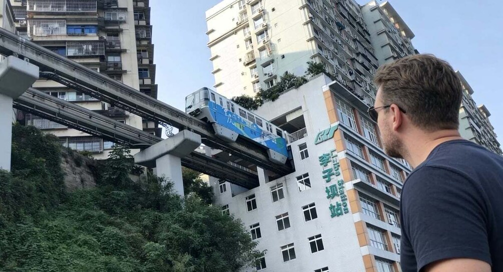 A person standing in front of a building with a blue train on an elevated track. 1