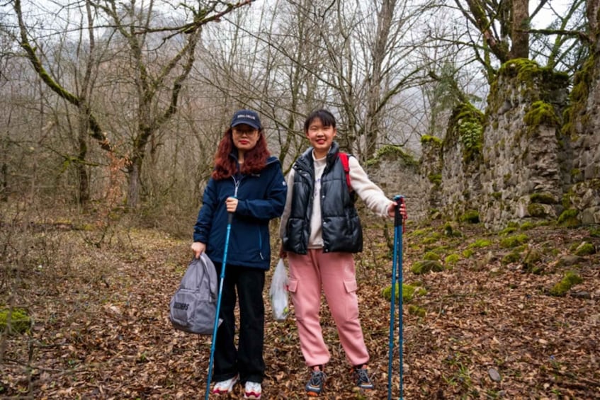 Two people hiking in a forest with leaf-covered ground and bare trees. 2