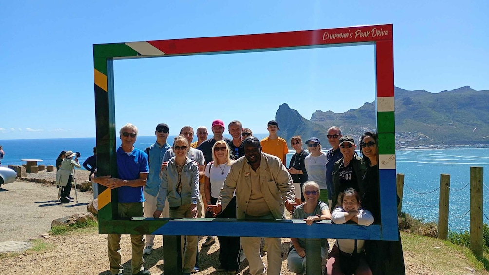 A group of people posing in front of a colorful frame with Chapman’s Peak Drive written on it, overlooking a coastal landscape with mountains in the distance. 1