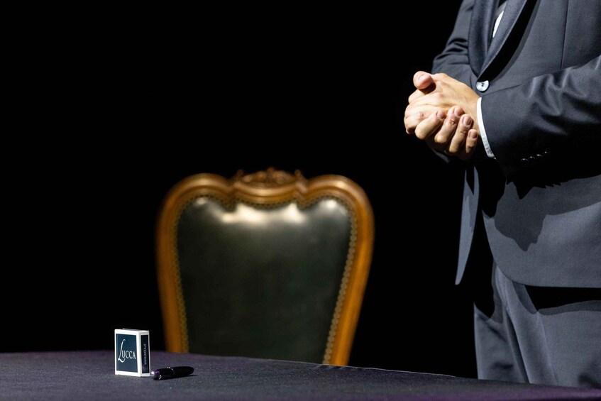 A person in a suit standing with hands clasped, a box labeled 'Lucca,' and a chair in the background. 3
