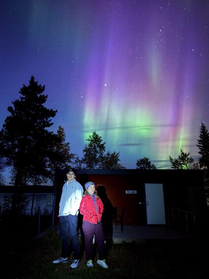 Deux personnes se tiennent devant un bâtiment sur lequel on peut lire l’inscription « MINILODGE ». En arrière-plan, un ciel nocturne étoilé et illuminé par des aurores boréales. 2