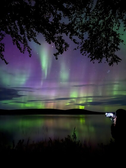 Des aurores boréales se reflètent sur un lac paisible, avec des silhouettes d’arbres et une personne tenant un appareil photo. 3