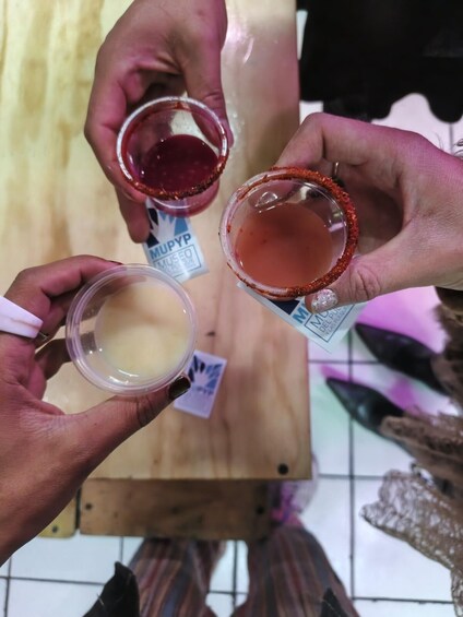 Three hands holding small containers with different colored liquids, possibly drinks, and a wooden surface in the background. 2