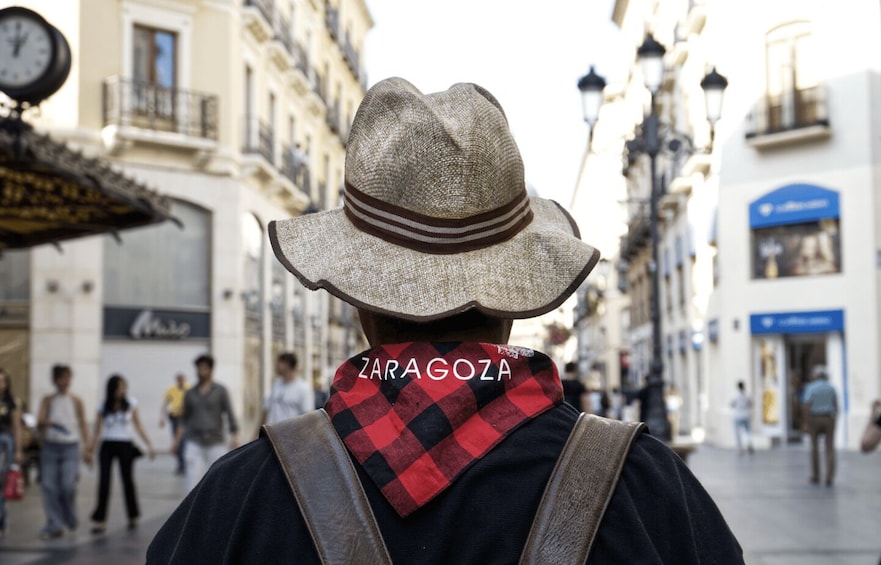 A person wearing a hat and scarf with 'ZARAGOZA' on it, walking on a street with buildings and shops. 5