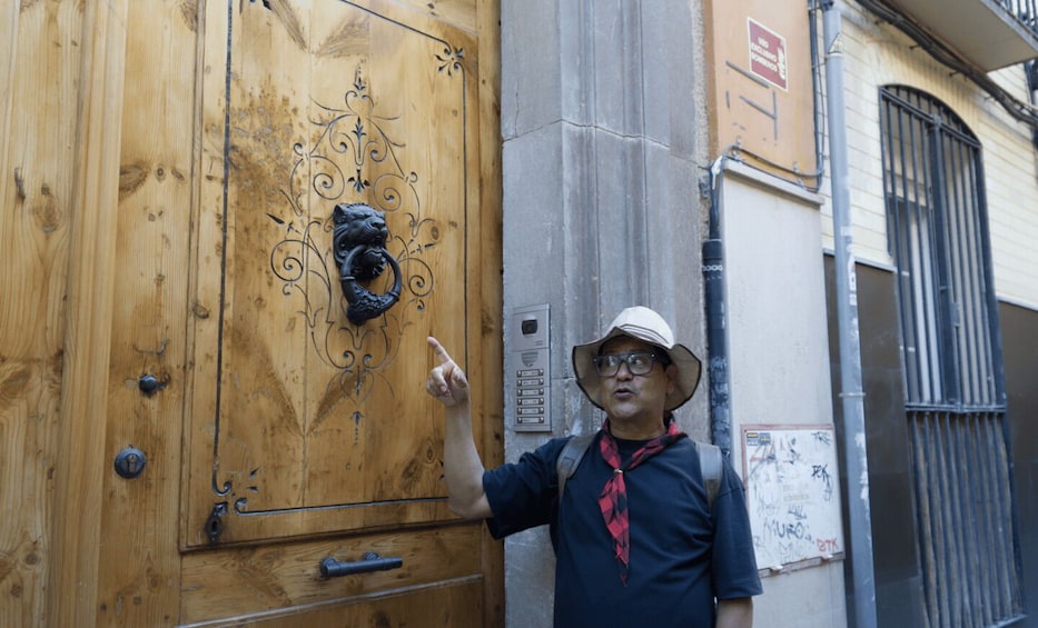 A person pointing at a wooden door with a lion head knocker, standing next to a building with a metal gate and a utility box. 3