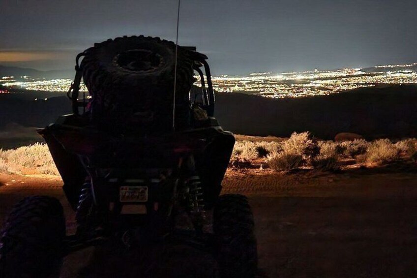 A vehicle with a large tire and a license plate, parked on a desert road with a cityscape in the background. 3