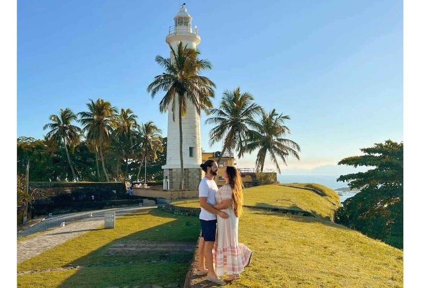 A couple standing in front of a lighthouse with palm trees and a coastal landscape. 3