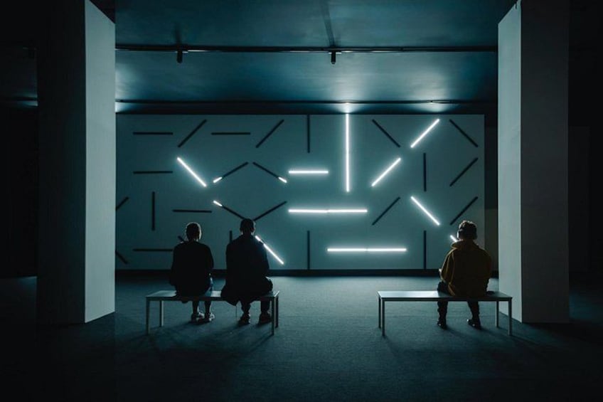 Three people sitting on benches facing a wall with geometric light patterns. 5
