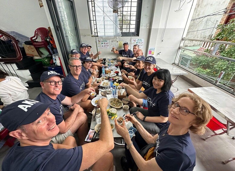 A group of people seated around a table, eating and drinking, in an indoor setting with a window and some posters on the wall. 3