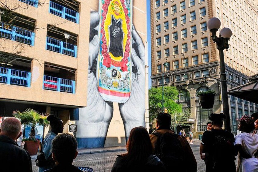 Un mural grande con manos sosteniendo un objeto colorido, y gente reunida alrededor. 3