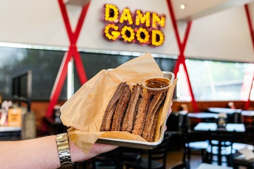 A tray of churros with a dipping sauce, held by a person wearing a watch, in a restaurant with a 'DAMN GOOD' sign in the background. 1