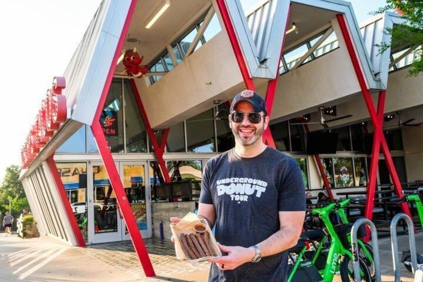 A person holding a donut in front of a building with a red and white facade and a sign that reads 'UNDERGROUND DONUT TOUR'. 3